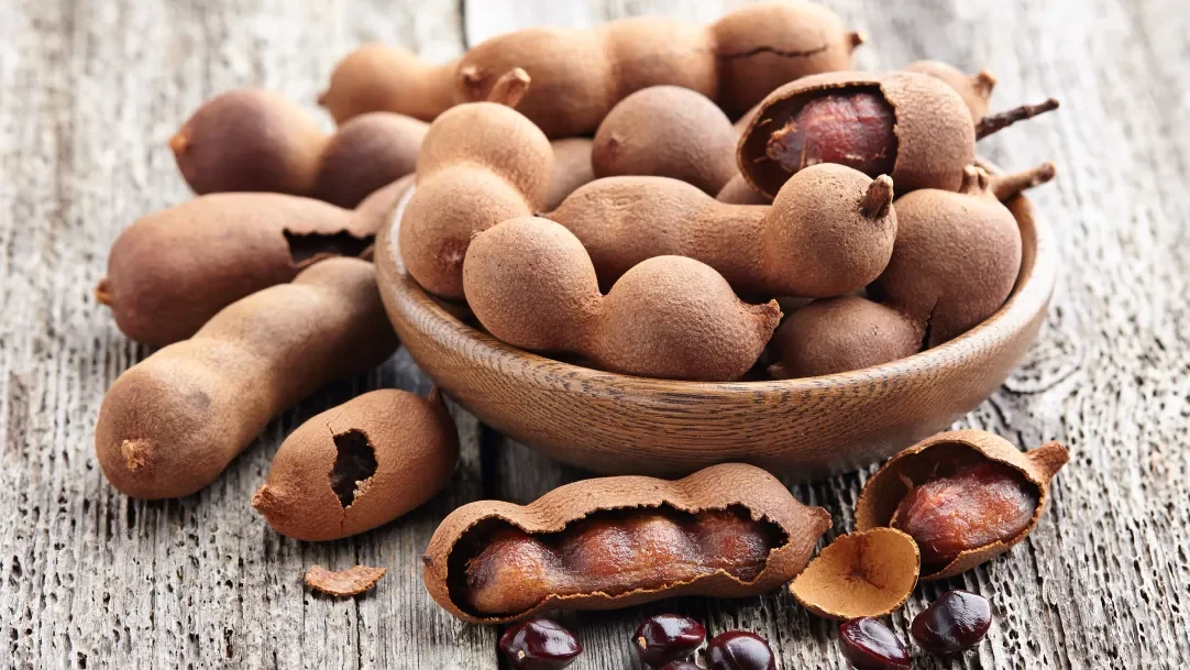 A wooden bowl filled with fresh tamarind pods, with some open pods showing the sticky pulp and seeds, placed on a rustic wooden surface