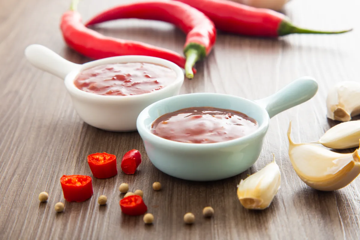 Two bowls of tamarind sauce surrounded by fresh garlic cloves, red chili peppers, and peppercorns on a wooden surface