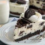 Close-up of a creamy Oreo Cheesecake slice with whipped cream and a mini Oreo cookie on a white plate.