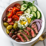 Mediterranean Steak Bowl with sliced steak, cucumbers, cherry tomatoes, olives, red onions, and yogurt sauce topped with olive oil.