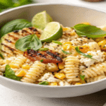 Creamy Street Corn Pasta Salad in a bowl with grilled corn, lime wedges, basil, and crumbled cheese
