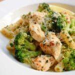 A close-up of baked lemon butter chicken with broccoli and pasta, topped with grated cheese and herbs.