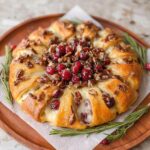 A golden-brown Brie & Cranberry Christmas Wreath Appetizer topped with pecans and fresh cranberries, garnished with rosemary.