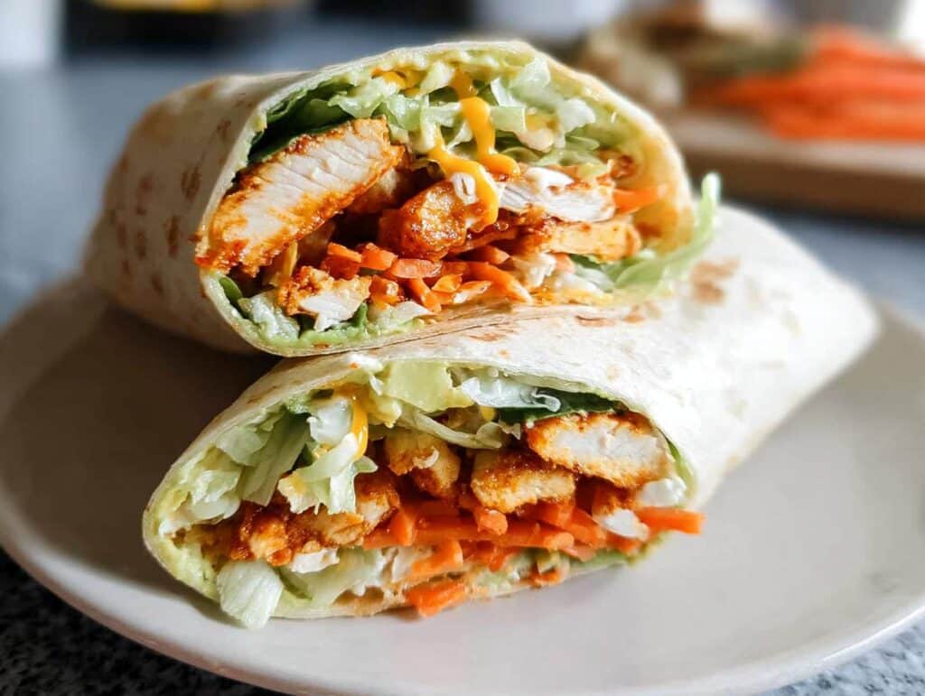 Close-up of two halves of a Buffalo Chicken Wrap filled with shredded lettuce, shredded carrots, and buffalo chicken pieces, drizzled with sauce.