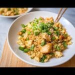 A close-up of a bowl of Chicken and Cauliflower Fried Rice, topped with green onions and sesame seeds, with chopsticks resting on the side.