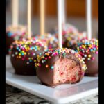 A close-up of a chocolate covered strawberry cake pop, with a bite taken out, revealing the pink cake inside and colorful sprinkles.