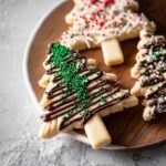 Close-up of decorated Christmas Shortbread Trees with chocolate drizzle and green sprinkles on a wooden plate.