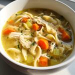 A close-up of a steaming bowl of Classic Chicken Noodle Soup, filled with noodles, chicken, carrots, and celery.