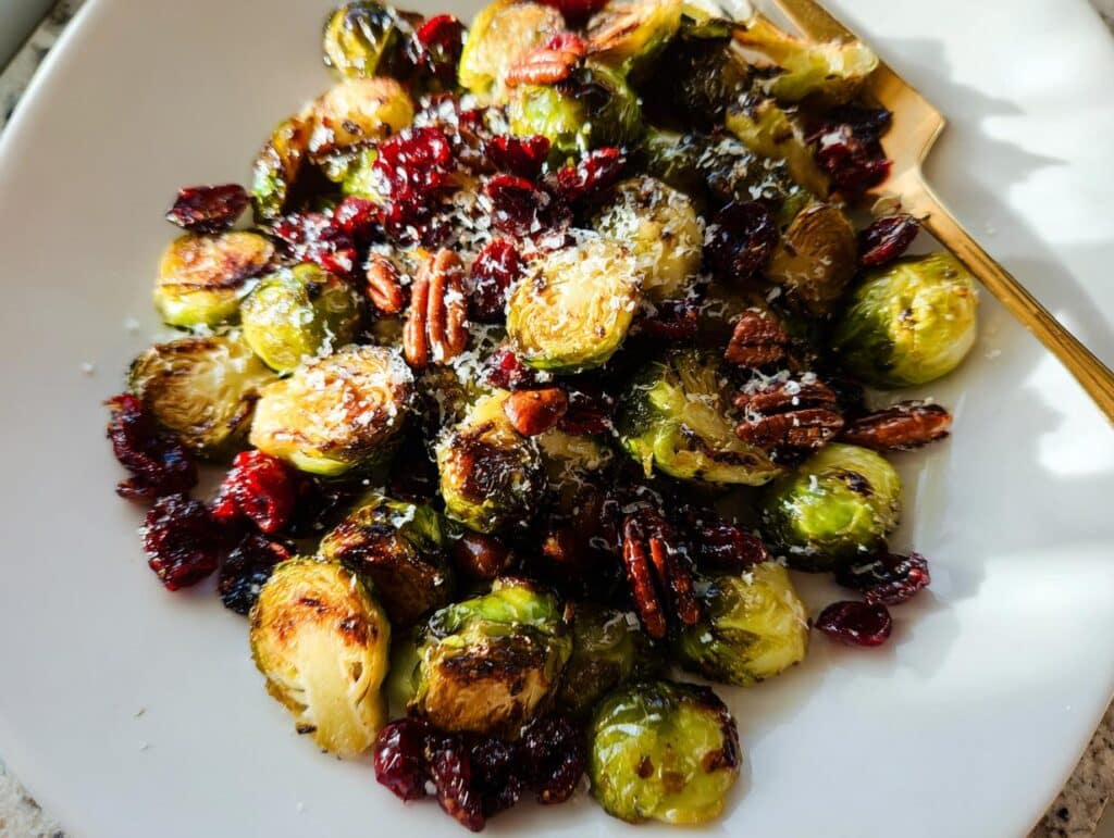 A white plate filled with roasted Cranberry Pecan Brussels Sprouts, garnished with shredded cheese and pecans.