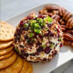 A festive Cranberry Pecan Goat Cheese Ball appetizer, covered in dried cranberries, chopped pecans, and green onions, served with crackers and whole pecans.