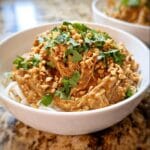 A bowl of Crockpot Thai Peanut Chicken served over white noodles, topped with chopped peanuts and cilantro.