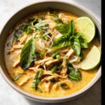 A close-up of a bowl of Easy Thai Chicken Curry Soup, featuring shredded chicken, rice noodles, fresh basil, and lime wedges.