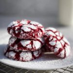 A stack of four Fudgy Red Velvet Crinkle Cookies dusted with powdered sugar on a white plate.