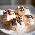 A close-up of several delicate Hot Cocoa Meringue Cookies topped with whipped cream and chocolate shavings.