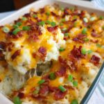 A serving spoon lifts a scoop of creamy Loaded Baked Potato Casserole, revealing melted cheese and crispy bacon.