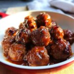 A close-up of a bowl filled with glistening Mini Cranberry Meatballs coated in a rich, glossy sauce with visible cranberry pieces and herbs.