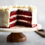 A slice cut from a decadent Red Velvet Layer Cake with Cream Cheese Frosting, revealing three rich red cake layers and white frosting.