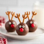 Three adorable Reindeer Cake Pops decorated with pretzel antlers, candy eyes, and red candy noses on a white plate.
