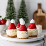 Four festive Strawberry Santa Cheesecake Cups topped with cream cheese frosting and a strawberry Santa hat.
