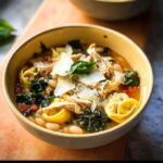 A close-up of a bowl of Tuscan Chicken Tortellini Soup, featuring tortellini, shredded chicken, white beans, kale, and shaved Parmesan.
