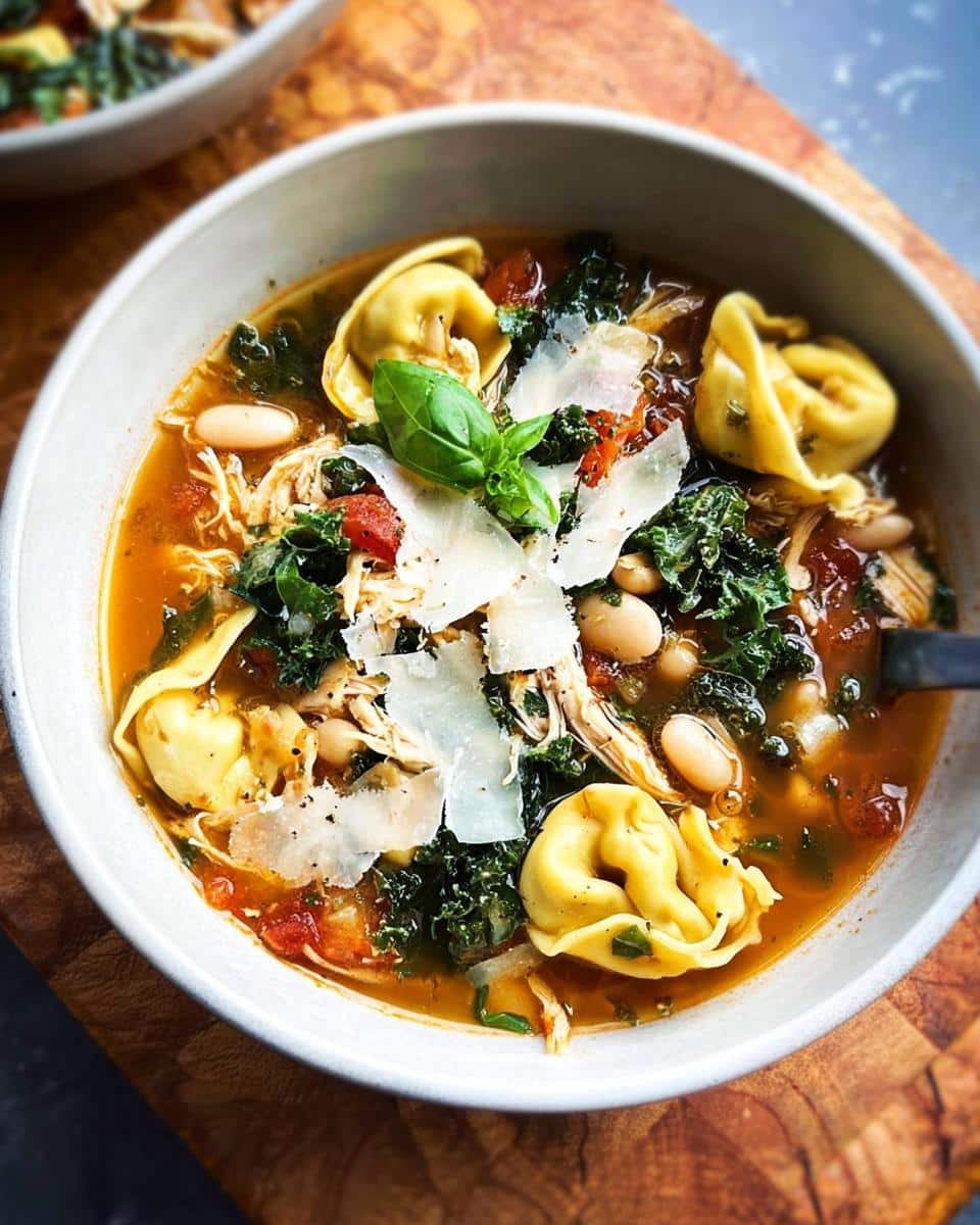A close-up of a bowl of Tuscan Chicken Tortellini Soup with tortellini, chicken, kale, beans, and shaved Parmesan.