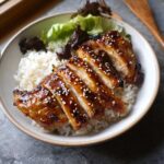 A bowl of white rice topped with sliced Air Fryer Teriyaki Chicken, drizzled with sauce and sprinkled with sesame seeds, served with lettuce.
