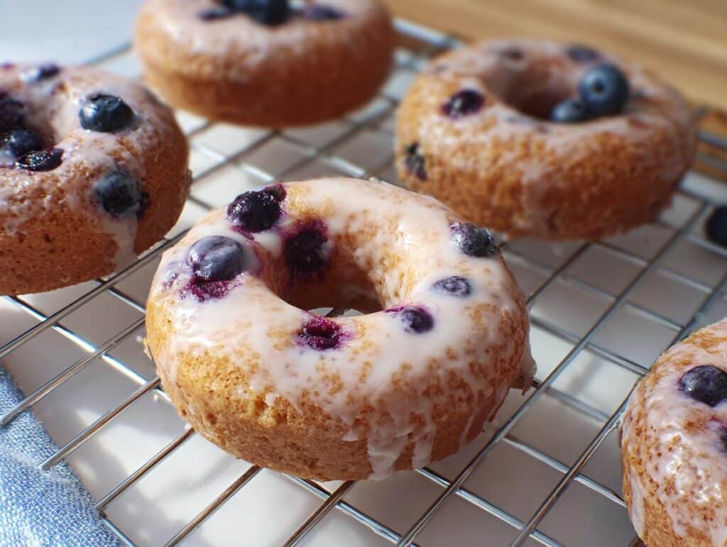 Close-up of several glazed Banana Blueberry Donut treats cooling on a wire rack, topped with fresh blueberries.