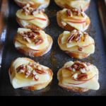 Close-up of several warm Brie, Apple, and Honey Crostini appetizers topped with pecans on a dark baking sheet.