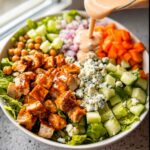 A close-up of Buffalo Chicken Salad Bowls being drizzled with dressing, featuring chicken, blue cheese, cucumber, and chickpeas.