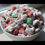 A white bowl filled with Christmas Puppy Chow coated in powdered sugar, featuring red and green colored pieces.