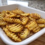 A close-up of golden, seasoned Crinkle-Cut Oven Chips piled high in a white serving dish.