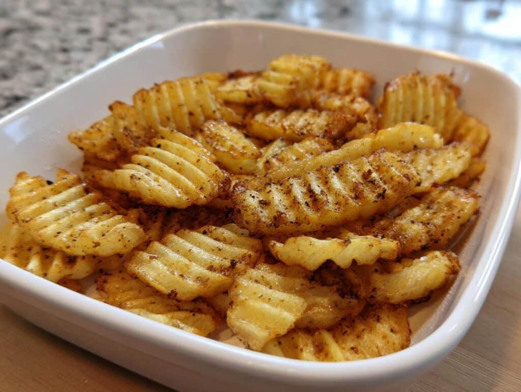 A close-up of golden, seasoned Crinkle-Cut Oven Chips piled high in a white square serving dish.
