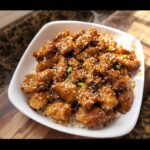 A bowl of Healthy Sesame Chicken served over white rice, garnished with sesame seeds and green onions, with steam rising.