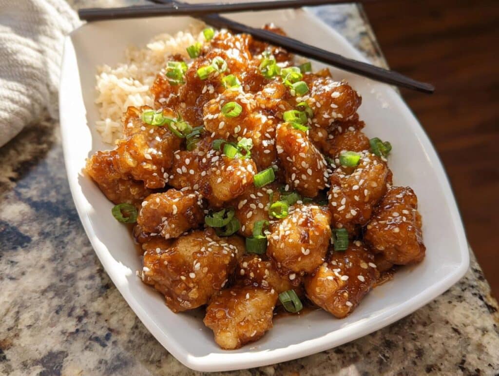 A plate of Healthy Sesame Chicken served with white rice and garnished with sesame seeds and chopped green onions.