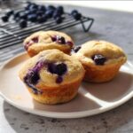 Three golden-brown High-Protein Blueberry Pancake Muffins with visible purple streaks from baked blueberries, sitting on a white plate.