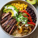 A vibrant High Protein Steak Fajita Bowl filled with sliced steak, black beans, corn, sautéed peppers and onions, avocado, and a drizzle of crema.