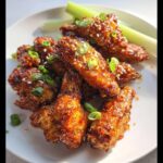 A plate of glistening Honey Garlic Air Fryer Chicken Wings, garnished with sesame seeds and chopped green onions, with celery sticks in the background.