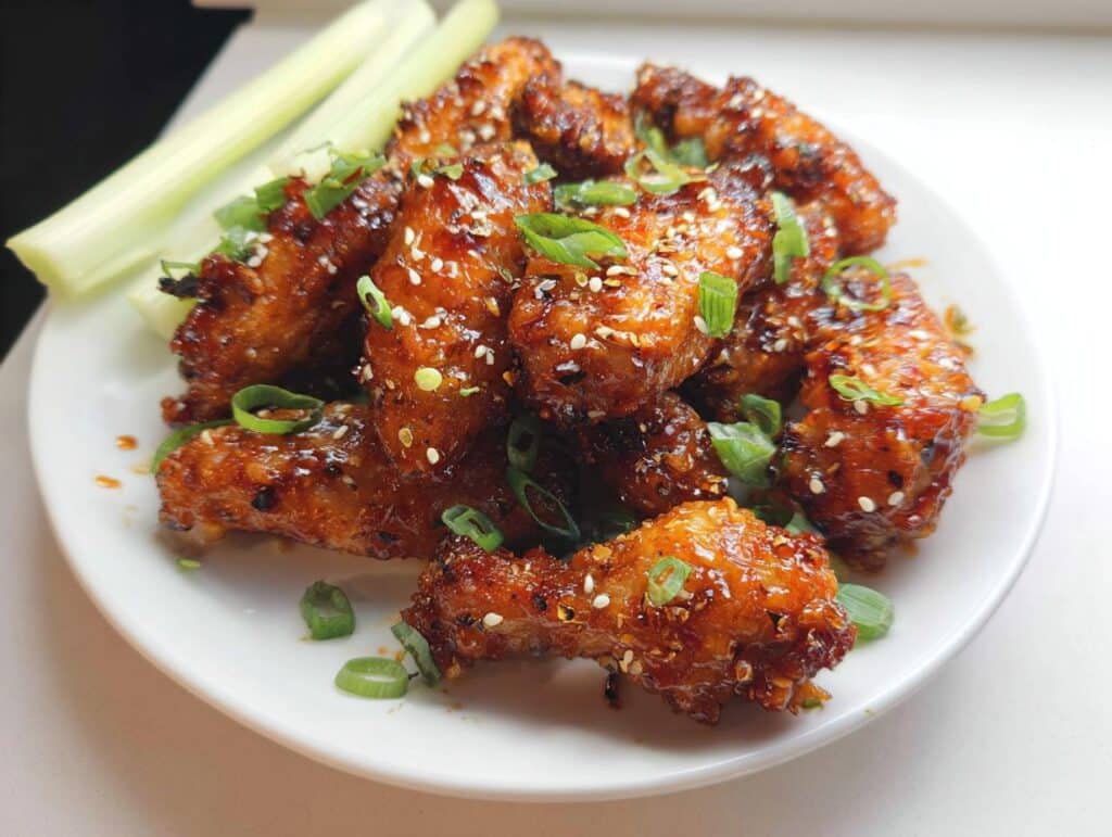 A plate of glistening Honey Garlic Air Fryer Chicken Wings, garnished with sesame seeds and chopped green onions, served with celery sticks.
