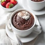 A rich Keto Chocolate Mug Cake topped with whipped cream and chocolate shavings, served in a white mug with a side of fresh strawberries.