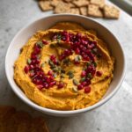 A bowl of bright orange Make-Ahead Pumpkin Hummus topped with ruby red pomegranate seeds and green pumpkin seeds.