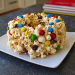 A ring-shaped Marshmallow Birthday Cake Popcorn treat studded with colorful M&M candies, resting on a white square plate.