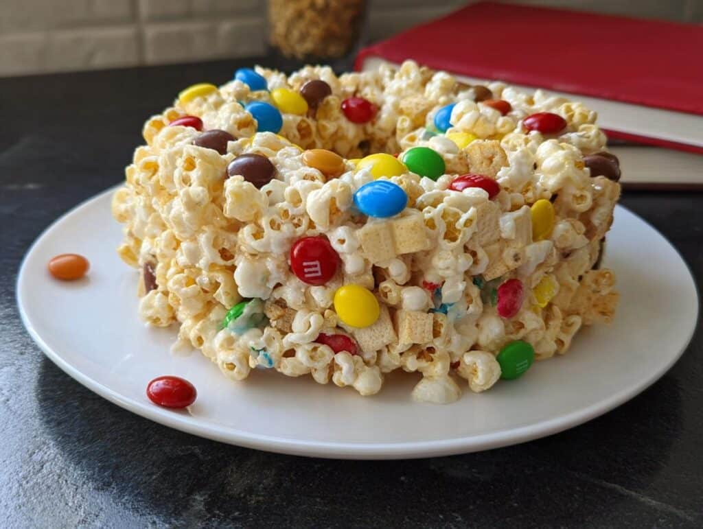 A molded ring shape of Marshmallow Birthday Cake Popcorn mixed with colorful candies on a white plate.