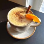 A close-up of a frothy Orange Cinnamon Milk Tea Latte in a mug, garnished with an orange slice and cinnamon stick.