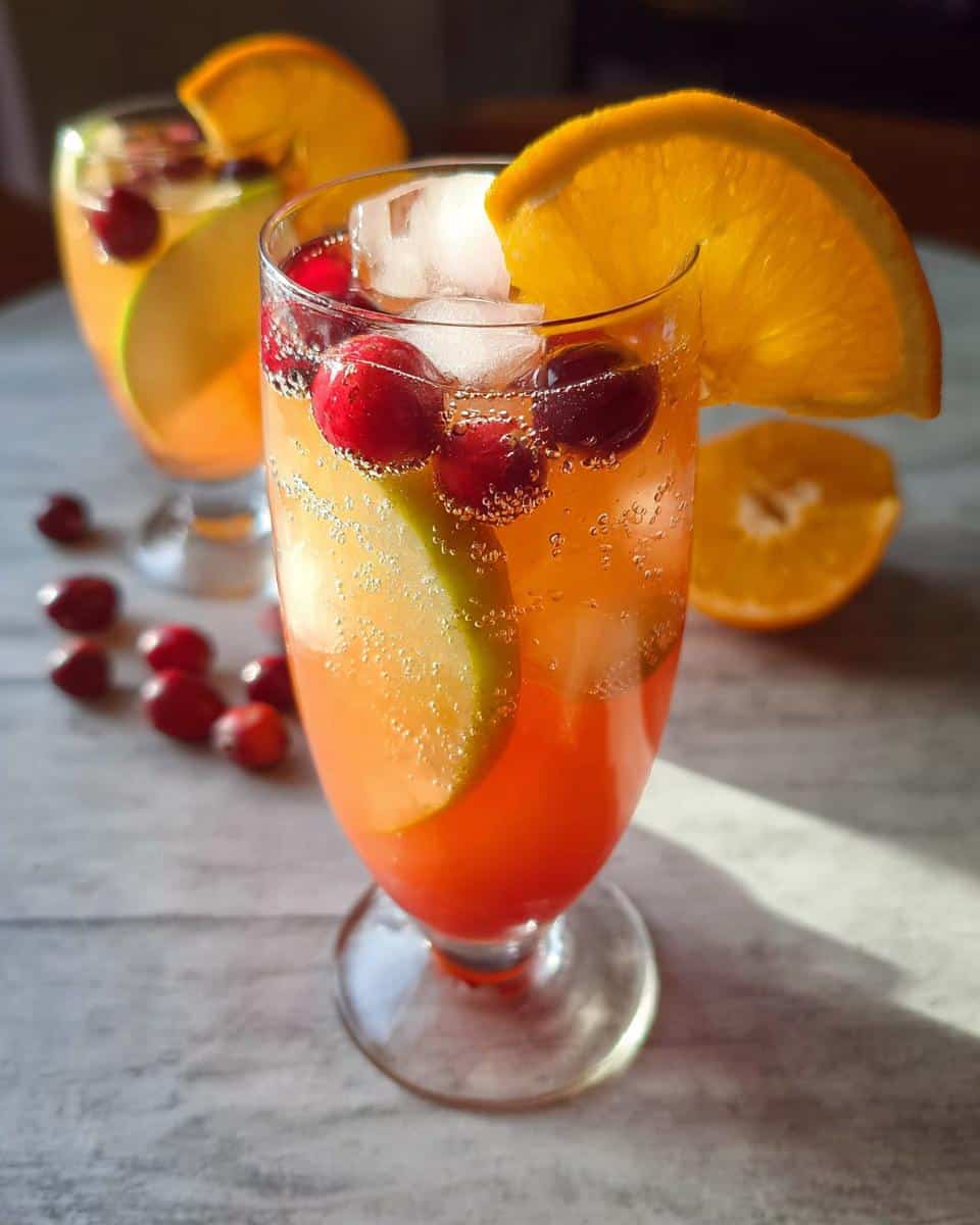 A close-up of a refreshing Orange Cranberry Sangria Mocktail with orange slices, cranberries, and apple slices in a glass, garnished with ice.