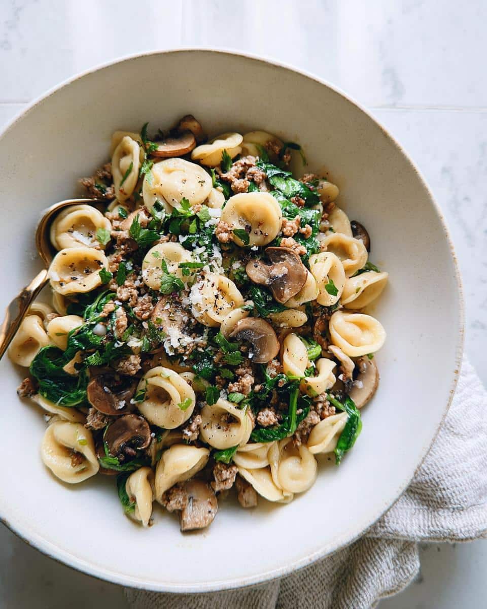 Close-up of orecchiette pasta mixed with ground turkey, sautéed spinach, mushrooms, and grated cheese in a light bowl.