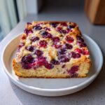 A square slice of Overnight Pancake Tray Bake topped with baked purple and red berries, served on a white plate.