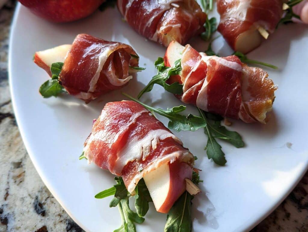 Close-up of several Prosciutto-Wrapped Pear Bites served on a white plate with arugula garnish.