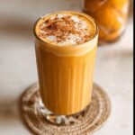 A tall glass of creamy Pumpkin Pie Smoothie Drink, topped with foam and a sprinkle of cinnamon.