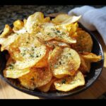 Close-up of crispy, golden Ranch-Seasoned Potato Chips dusted with seasoning and parsley in a dark bowl.