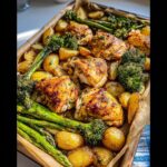 A close-up of a sheet pan filled with Sheet Pan Garlic Butter Chicken & Veggies, including roasted chicken thighs, potatoes, broccoli, and asparagus.